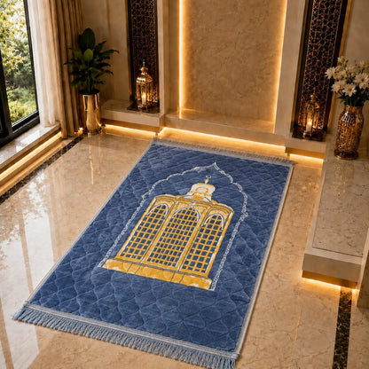 A blue rectangular prayer rug with a gold and white mosque design and fringe, displayed on a polished marble floor in a luxurious, warmly lit interior setting.