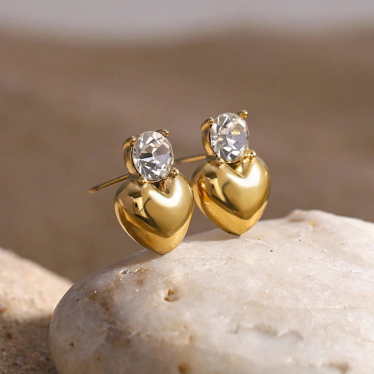 Gold heart-shaped earrings with clear gemstones on a stone surface